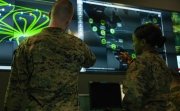 Two soldiers have their back to the camera while looking at a green and blue display of digital information.