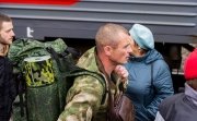 A white man in green camoflage, wearing a green camoflage backpack, walks in front of a train. 