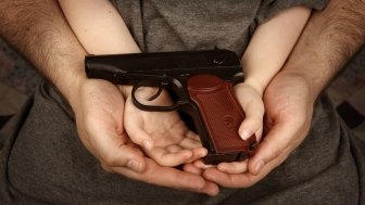 A man cradles a boy's hands while holding a gun.