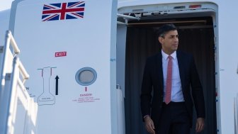 Prime Minister Rishi Sunak exits a plane with a UK flag visible on the door.