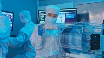 A woman in a clean suit holds up a silicon wafer in a laboratory.