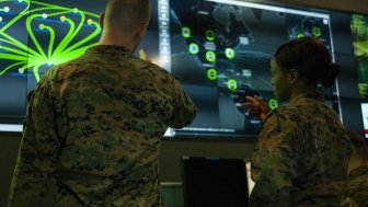 Two soldiers have their back to the camera while looking at a green and blue display of digital information.