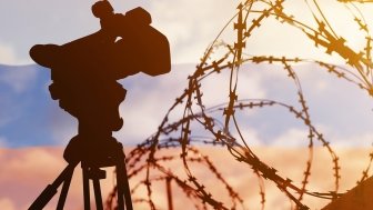 A composite photo with the Russian flag in the background, and barbed wire and a television news camera in the foreground