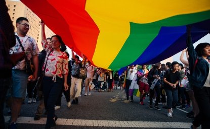 A group of people march carrying a large rainbow flag above their heads.